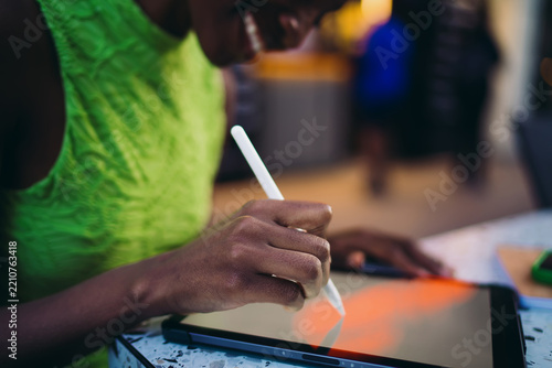 Close up of dark skinned female hand drawing on touch screen tablet with digital stylus pen. Smiling African American artist creating sketch using professional software at cafe.