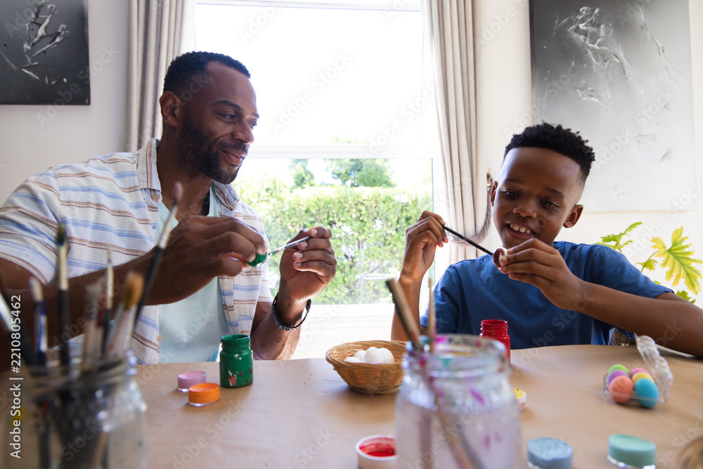 Obraz premium African American father and son painting eggs at table by window with paint pots