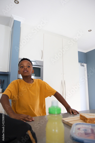 African boy in mustard tee standing with hands on kitchen counter near sandwich and water bottle