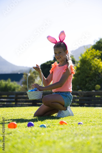 Girl child kneeling on lawn, holding white basket with colorful eggs and wearing bunny headband