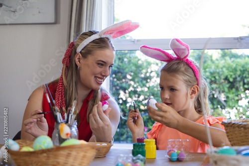 Mother and daughter sitting at home table painting eggs with brushes wearing bunny ear headbands