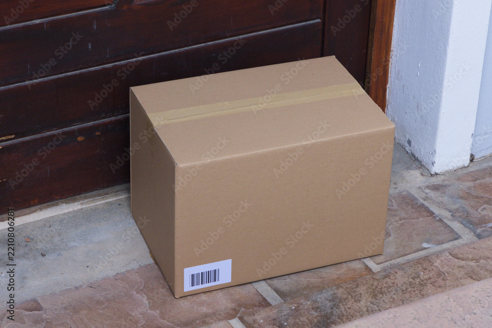 Fototapeta premium Sealed cardboard box is sitting on tiled threshold near dark wooden door with barcode label