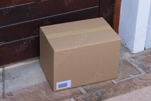 Sealed cardboard box is sitting on tiled threshold near dark wooden door with barcode label