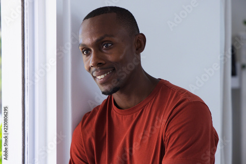 African American man leaning against pale wall near window, looking right, wearing rust-red t-shirt