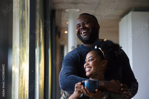 African American couple standing, embracing near tall glass windows in corridor holding blue mug