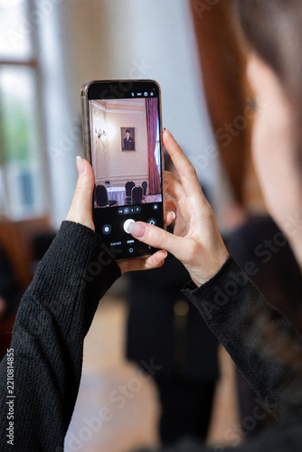 Woman holding a smartphone to take a photo of an elegant interior hall with classic decor and painting. Female tourist capturing historical building architecture using her mobile phone.