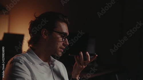 Closeup face of cheerful Caucasian man with glasses recording voice note on personal device sitting at desk, happily communicating during overtime work in cozy, dark office interior, slow motion.