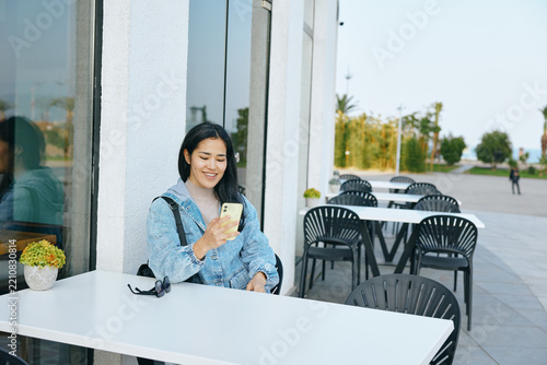 Woman outdoor cafe seating at a white table, smartphone in hand, casual relaxed mood, urban daylight, showcasing technology and connection during a calm moment