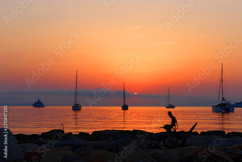 Sailboats and yachts silhouetted at sunset over calm Mediterranean waters with rocky shore, Antibes.