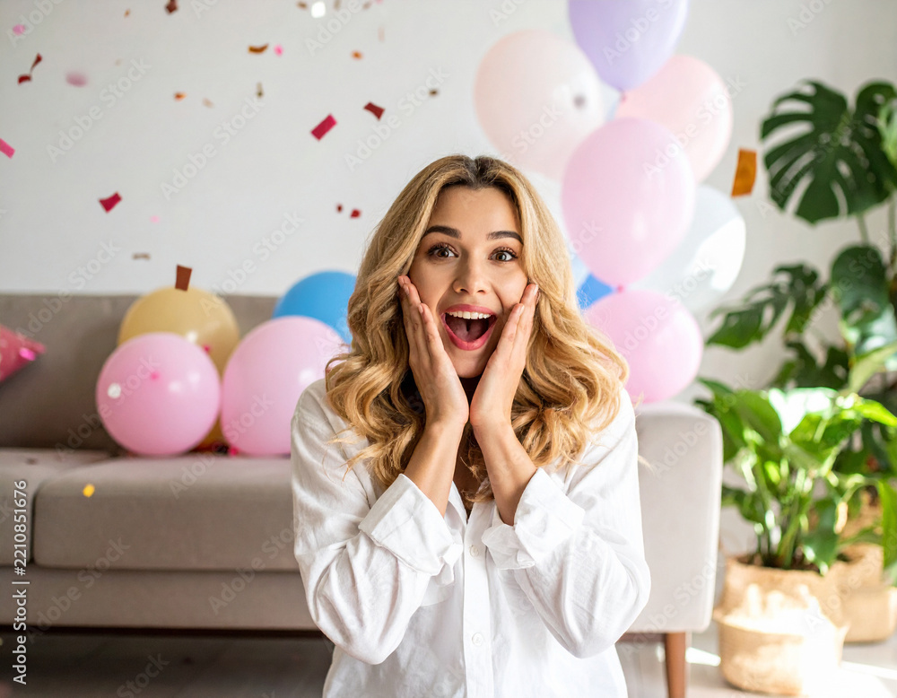 Fototapeta premium A joyful woman experiences a burst of excitement, hands on cheeks, surrounded by vibrant balloons and festive confetti. This celebration captures pure delight for birthdays or special surprises.