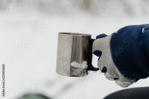 Gloved hand holding a metal mug on a cold winter day outdoors. Winter glove gripping a thermal cup for a hot beverage in snowy weather. Cozy comfort of a warm drink during a frosty outdoor adventure