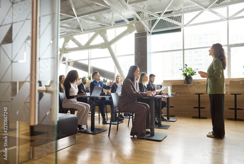 Speaker leads training seminar for office team. Engaged audience in a meeting listens to a coach giving a presentation in a bright modern workspace. Clear concept of team learning workshop.