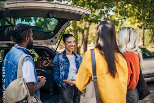 A group of diverse friends stands by an open car trunk, chatting and laughing as they prepare for a road trip. The warm sunlight filters through the trees, creating a joyful atmosphere for their adven