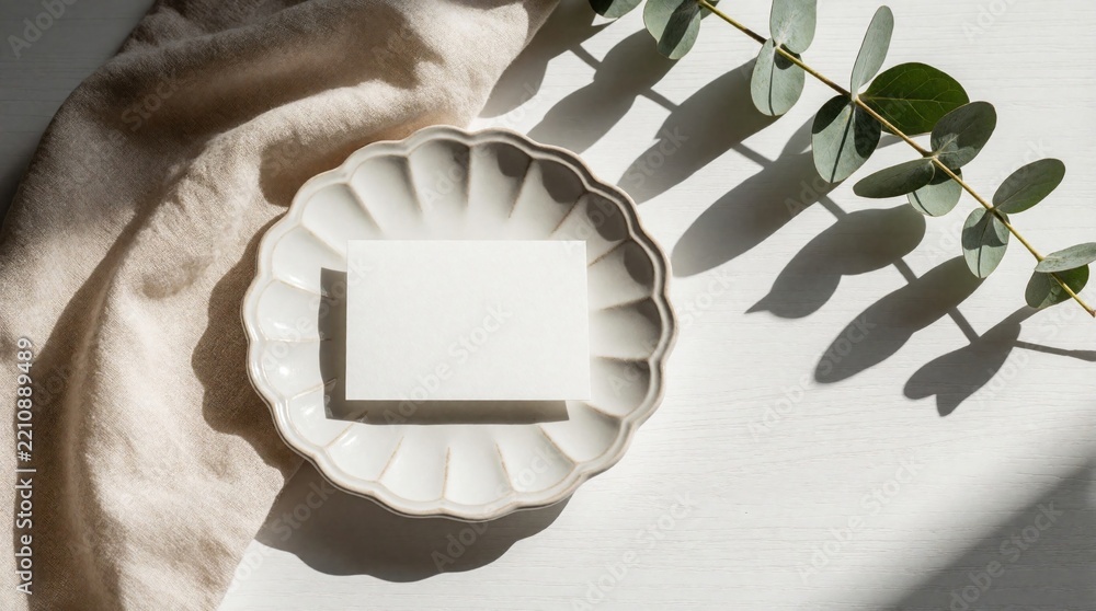 Naklejka premium Minimalist mockup features blank white card on scalloped ceramic plate. Eucalyptus branch, beige linen fabric rest on white wooden table. Natural sunlight creates soft shadows for wedding invitation