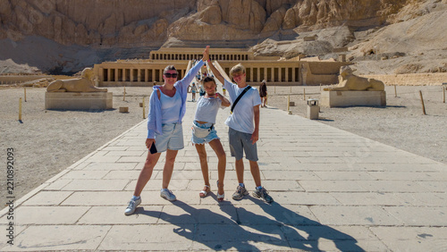 Family of three posing for photo in front of Mortuary Temple of Hatshepsut in Egypt, with sphinx statues visible.