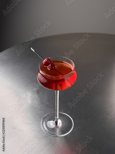 Red cocktail in coupe glass with cherry garnish on stainless steel table, studio lighting closeup showing glossy reflections and rich crimson color elegant beverage