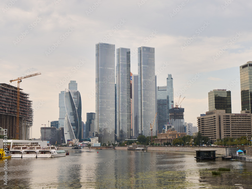 Fototapeta premium Modern city skyline with glass skyscrapers along riverfront, construction cranes at building site, boats docked near promenade, and overcast clouds