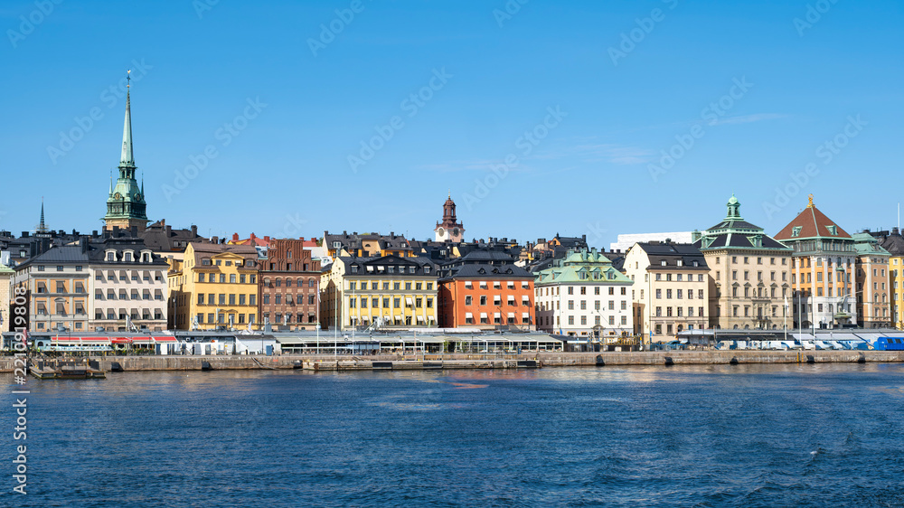 Fototapeta premium Panoramic view of colorful historic buildings, German Church spire, and waterfront promenade along Skeppsbron in Gamla Stan under a clear blue sky in Stockholm