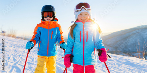 Two smiling children dressed in colorful ski attire, with helmets and goggles, posing on a snowy mountain slope with ski poles in hand