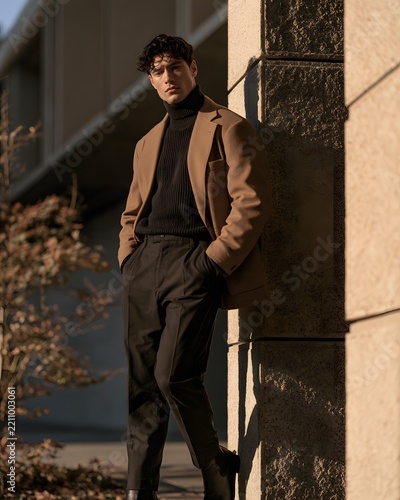 Stylish man in a beige blazer and black turtleneck leaning against a stone wall during golden hour