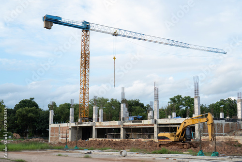 Tall crane standing above active construction site with yellow excavator machinery working on building structure showcases ambitious industry development under blue sky