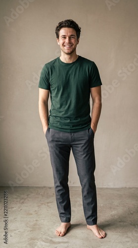 Full length portrait of a smiling young man standing barefoot in a green tshirt and grey trousers against a neutral studio background