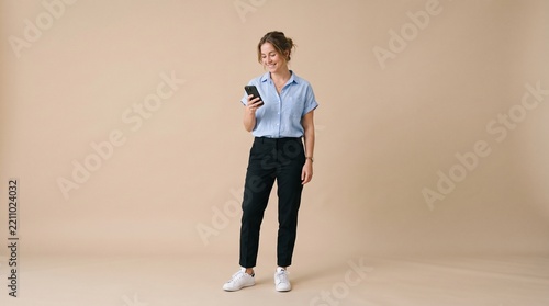 Full length portrait of a smiling young woman standing against a neutral beige background while looking down at her smartphone in her hand