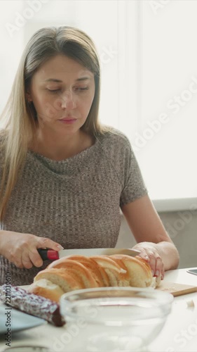 Woman carefully slices loaf of bread on wooden cutting board in soft natural light. scene includes additional food items like chocolate and glassware with focus remaining steadily on her