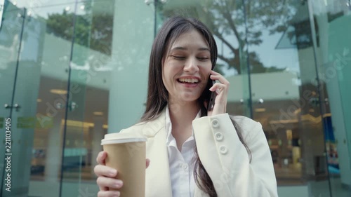 Medium close-up portrait of a successful young Asian businesswoman wearing a white suit having a pleasant mobile phone conversation outdoors near a commercial building. Concept of client communication