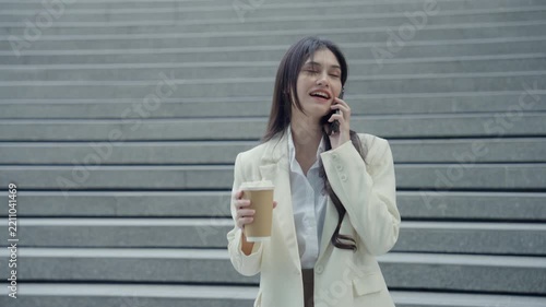Medium close-up portrait of a successful young Asian businesswoman wearing a white suit having a pleasant mobile phone conversation outdoors near a commercial building. Concept of client communication