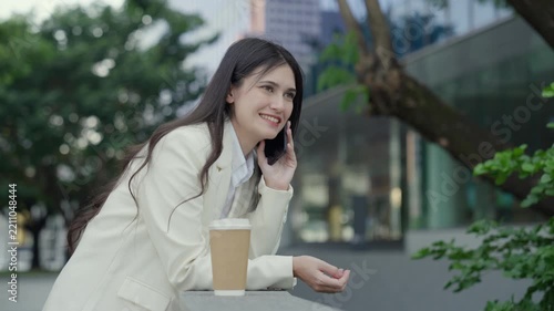Medium close-up portrait of a successful young Asian businesswoman wearing a white suit having a pleasant mobile phone conversation outdoors near a commercial building. Concept of client communication