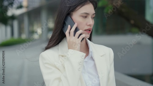 Medium close-up portrait of a successful young Asian businesswoman wearing a white suit having a pleasant mobile phone conversation outdoors near a commercial building. Concept of client communication