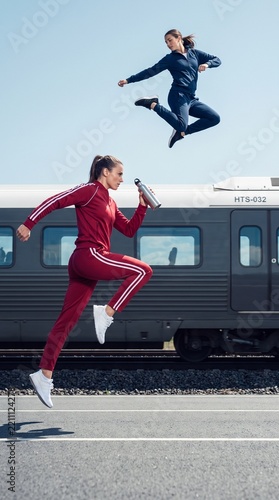 Two athletic women jumping in tracksuits beside a train
