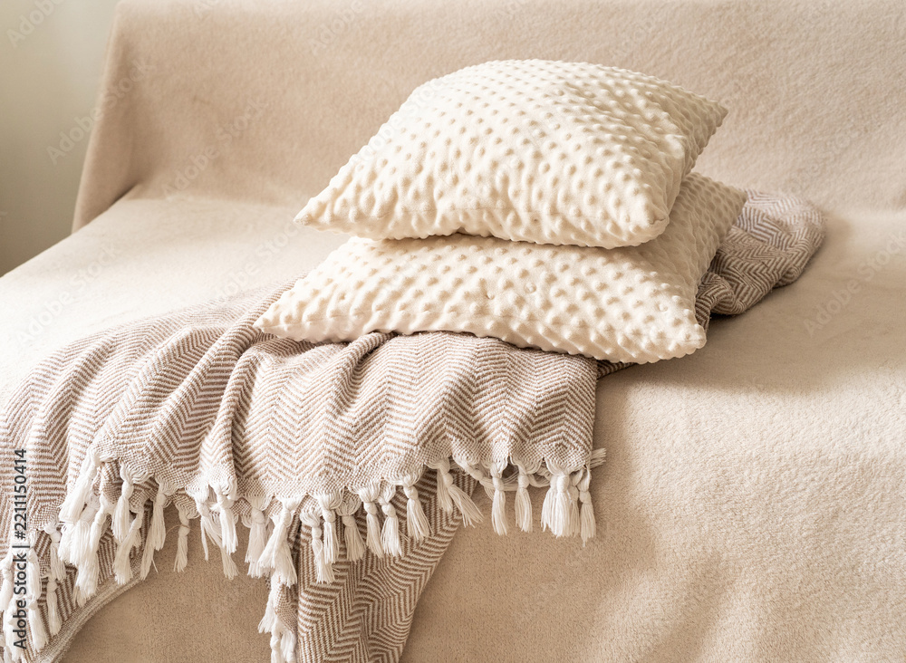 Obraz premium Textured decorative pillows in milky-beige hue, stacked atop patterned throw blanket with white fringe, resting on beige sofa against neutral backdrop. Quiet luxury, cozy home accessories