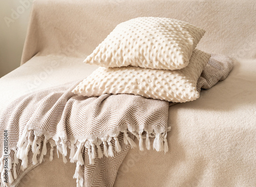Textured decorative pillows in milky-beige hue, stacked atop patterned throw blanket with white fringe, resting on beige sofa against neutral backdrop. Quiet luxury, cozy home accessories
