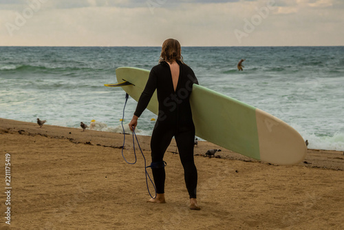 Fotografie Surfer carrying surfboard and wearing wetsuit walking toward ocean waves on sand