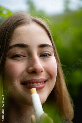 Fotografie Close-up portrait of a smiling woman using a lip balm stick outdoors, demonstrating everyday lip care and skincare, surrounded by green foliage in nature