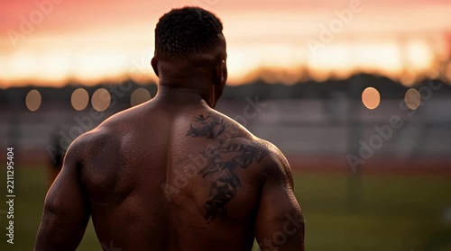 Athletic Man Training Outdoors at Sunset; Powerful Back Muscles and Tattoos Visible During Intense Workout on Track Field with Vibrant Orange Sky Backdrop