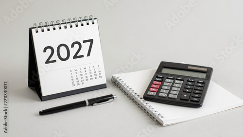 2027 desktop calendar with a black calculator and a pen on a notebook on a grey surface with calendar stands upright next black electronic spiral bound notebook year