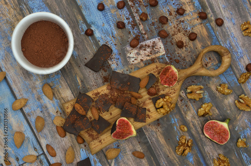Dark chocolate squares, cocoa powder in bowl, nuts, almonds and fig on dark blue boards. Preparing to make an autumn or winter chocolate dessert or hot drink.