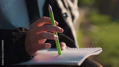 Close up view shows person hands resting on opened spiral bound notebook. green pen is held in one hand poised above paper which contains faint handwritten notes and grid lines. Soft