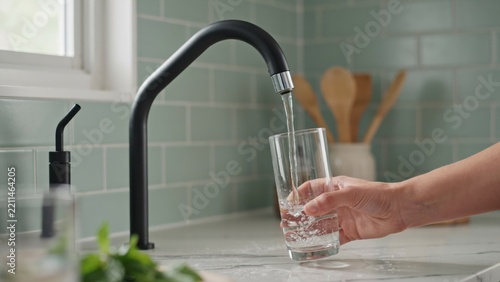 A hand fills a clear glass with fresh, clean water from a sleek black kitchen faucet in a bright modern kitchen. Perfect for health, wellness, and sustainable living themes.