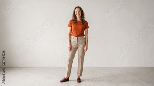 Full body portrait of a smiling young woman standing against a neutral wall, wearing a rustcolored tshirt, beige trousers, and brown leather loafers