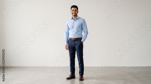 Full body portrait of a young professional man standing confidently in a light blue buttondown shirt and navy blue dress pants against a plain wall