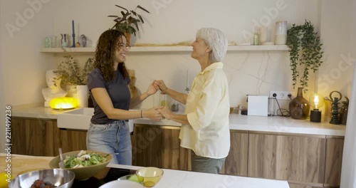 Cheerful mother and her adult daughter dancing and having fun while preparing a fresh vegetable salad