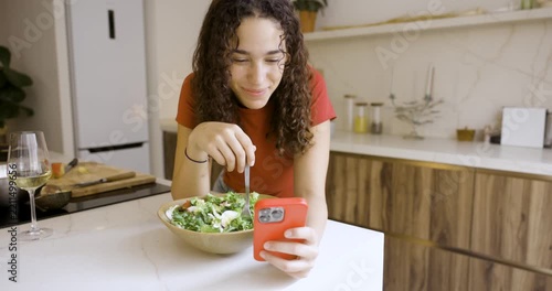 Young woman with curly hair having a video call on her phone while eating a fresh salad in her kitchen