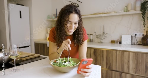 Happy young woman eating a healthy salad and browsing social media on her smartphone in a modern kitchen