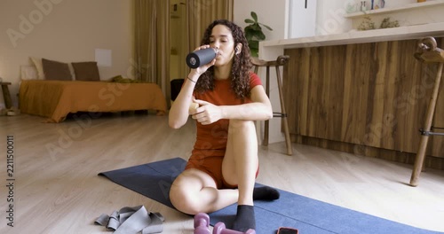 Athletic young woman sitting on yoga mat taking a break, drinking water and using her smartphone
