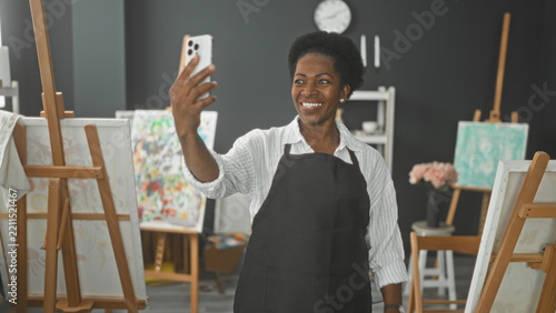 Obraz na plátně Woman painter in apron holds smartphone at arm's length, smiling and waving hand in studio; creative joy