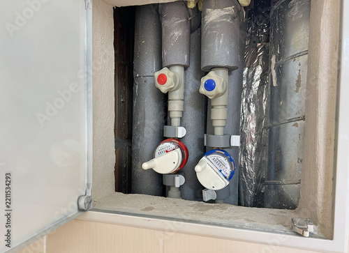 Hot and cold water meters with IoT remote reading modules, valves and insulated pipes installed inside a residential plumbing service cabinet.